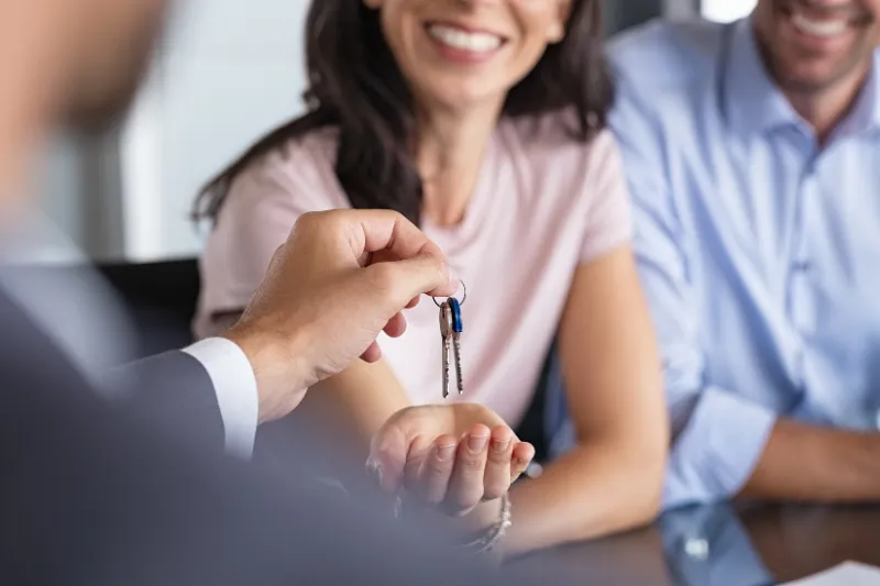 A person in a suit hands house keys to a smiling woman, who is reaching out her hand to receive them, while another person sits beside her, both appearing happy.
